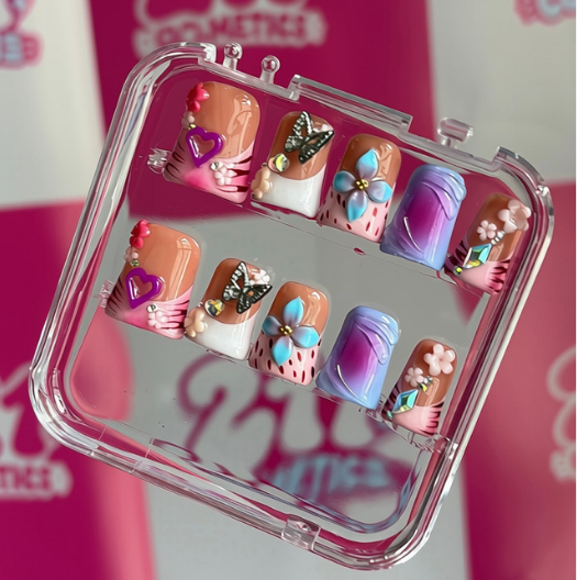 A clear plastic case holding six decorative nail wraps or false nails with colorful floral and abstract designs, photographed against a pink-and-white checkered branded backdrop.