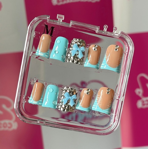 A set of false press-on nails in a clear plastic case, featuring pastel blue, peach, and silver glitter designs, displayed against a pink and white checkered background with 'MS Cosmetics' logos.